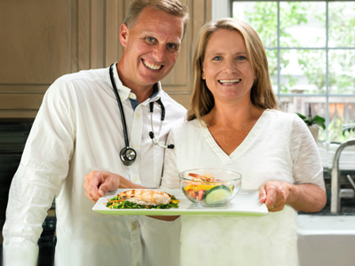 A doctor and patient standing in front of the camera holding a tray of healthy food. 